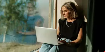 Ung kvinna jobbar på en laptop. Hon är i hemmiljö och lutar sig mot en vägg. Det är en stor fönsterruta där man kan se grönska och träd. I bakgrunden skymtar vatten. 