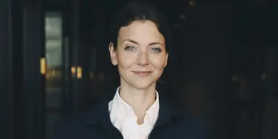 Ung och välklädd kvinna står med händerna i fickorna och tittar rakt in i kameran. Det är en mörk bakgrund. Hon har ett svagt leende och upplevs självsäker.