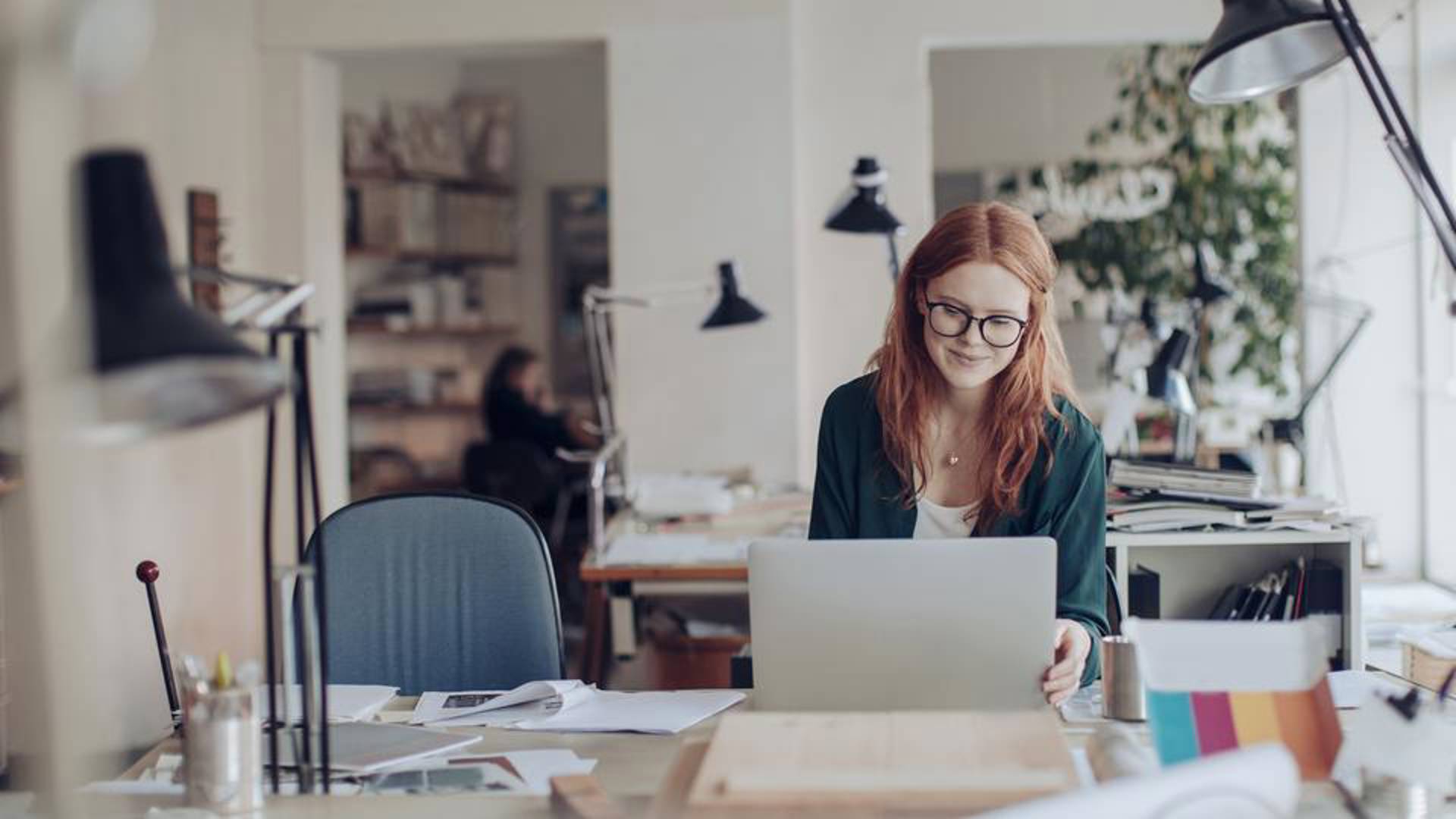 Ung kvinna som arbetar framför dator i kontorsmiljö med person i bakgrunden