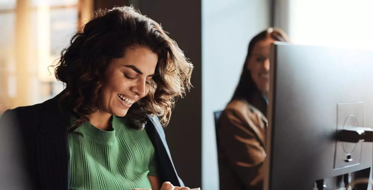 Kvinna med brunt lockigt hår och grön tröja med kavaj om axlarna är i fokus i bilden. Hon håller i en mobil med båda händerna och tittar glatt på skärmen. Det sitter en kvinnlig kollega till höger om henne och det är på en arbetsplats. Det är en dataskärm framför på skrivbordet. 