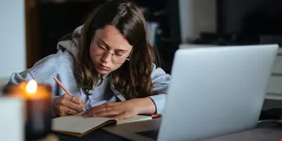 Ung student med glasögon sitter hemma och skriver koncentrerat i en anteckningsbok. Det står ett tänt ljus bredvid en laptop. 