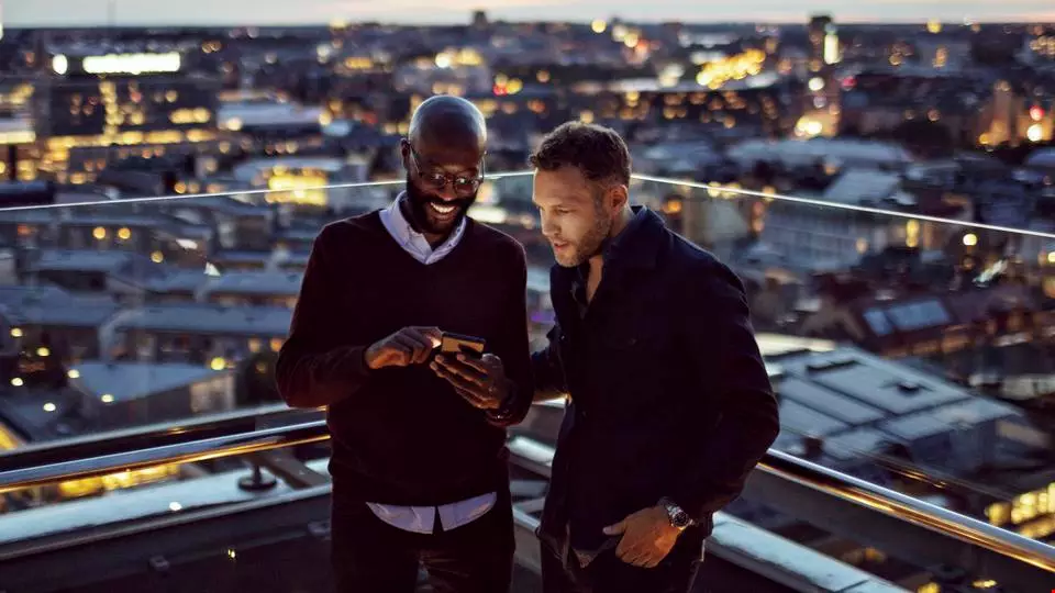 Två män står bredvid varandra och tittar på något som de ser på en mobilskärm. De står utomhus med utsikt över en stad under en varm sommarkväll. 