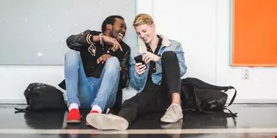 Två unga män sitter på golvet i en korridor och skrattar åt något på den ena killens mobilskärm