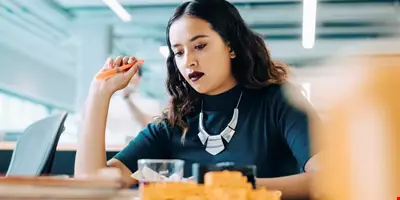 En ung kvinna med mörkt läppstift  håller i en orange penna och tittar fokuserat på något framför sig på bordet. Det är lysrör i taket och man får en minimalistisk och kreativ känsla av arbetsplatsen. 