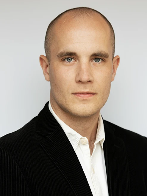 Man med vit skjorta, svart kavaj, rakat huvud ser bestämd ut.