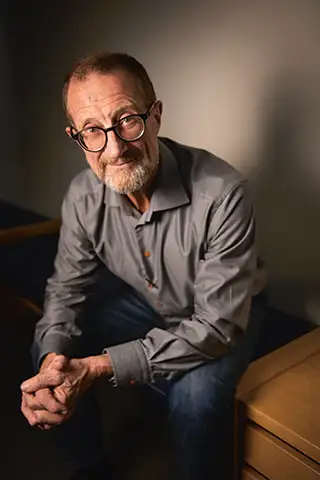 Man i medelåldern, han har skägg, glasögon med bred båge, är klädd i jeans och skjorta. Sitter lutad framåt.