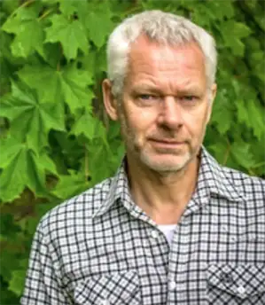 Man med kort grått hår och rutig skjorta, står framför ett lövverk.